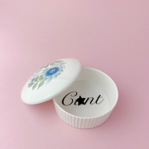 May include: White ceramic trinket box with a floral design on the lid. The box is open and the inside says "Cant" with a star.