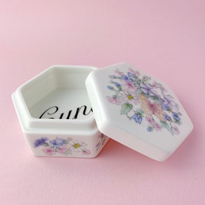 May include: A white, hexagonal trinket box with a floral design in shades of pink, purple, and blue. The lid is slightly ajar, revealing the interior and the word "curio" in black script. The box sits on a pink surface.
