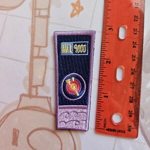 May include: Purple and blue embroidered patch with the text "HAL 9000" in yellow. The patch features a stylized image of a red and yellow target with a white arrow.