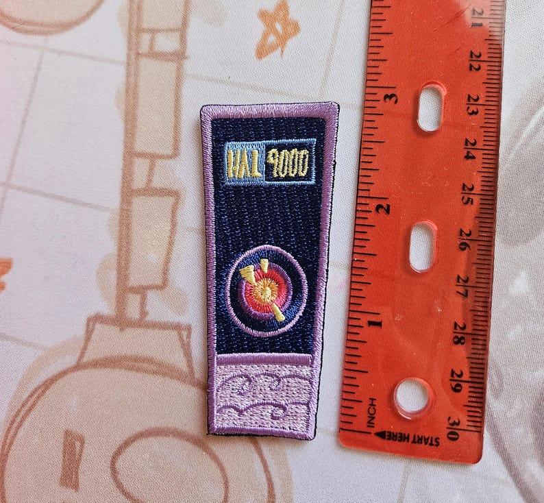 May include: Purple and blue embroidered patch with the text "HAL 9000" in yellow. The patch features a stylized image of a red and yellow target with a white arrow.