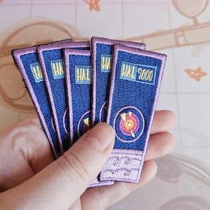 May include: Five blue and purple embroidered patches with the text "HAL 9000" on the front. The patches have a pink border and a yellow and red target design.