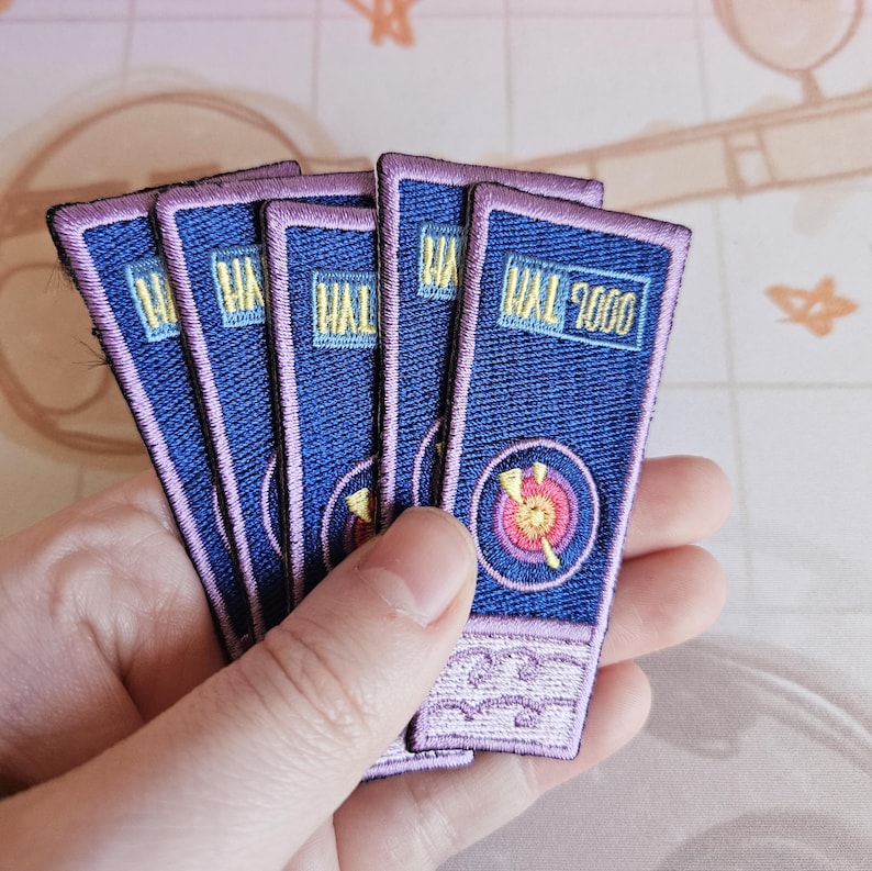 May include: Five blue and purple embroidered patches with the text "HAL 9000" on the front. The patches have a pink border and a yellow and red target design.