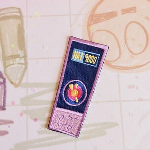Peut inclure: Un patch brodé rose et bleu avec le texte "HAL 9000" et un motif circulaire rouge, orange et jaune.