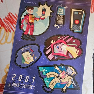 May include: A set of five stickers featuring characters and objects from the film "2001: A Space Odyssey". The stickers are on a blue background with stars. The stickers include a red space suit, a HAL 9000 computer, a red and white spaceship, a notepad with a pencil, and a spaceman floating in space.