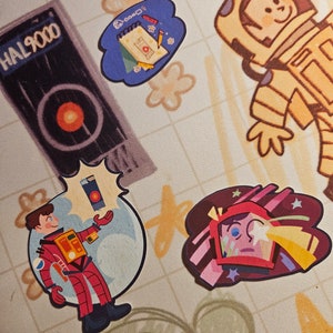 May include: Four colourful stickers featuring illustrations of an astronaut, a notebook, a spaceship, and a person in a red spacesuit. The stickers are on a beige background with a grid pattern.
