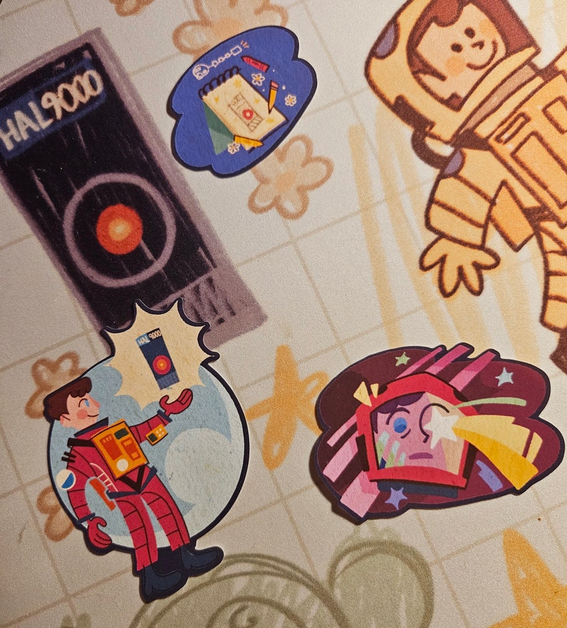 May include: Four colourful stickers featuring illustrations of an astronaut, a notebook, a spaceship, and a person in a red spacesuit. The stickers are on a beige background with a grid pattern.