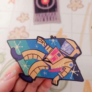 May include: A yellow astronaut sticker with a blue, green, and yellow space background. The astronaut is wearing a helmet and has a pink and blue space suit. The sticker has a retro, 1950s style.