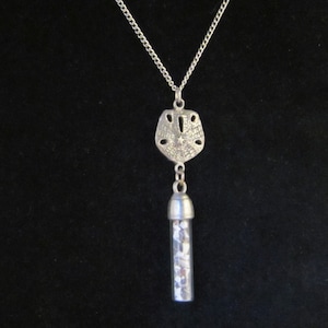 May include: A silver necklace with a silver pendant shaped like a seashell. The pendant has a small glass vial filled with white sand hanging from it.