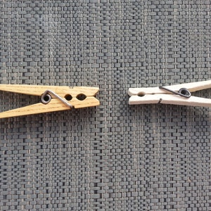May include: Two wooden clothespins with metal springs. One is natural wood, the other is painted white. The clothespins are on a textured gray surface. These are useful for hanging laundry or crafts.