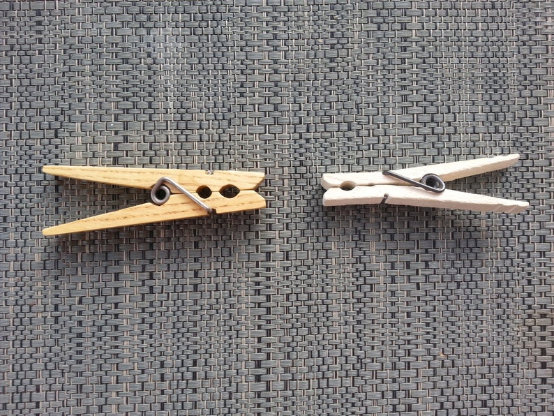 May include: Two wooden clothespins with metal springs. One is natural wood, the other is painted white. The clothespins are on a textured gray surface. These are useful for hanging laundry or crafts.