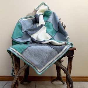 May include: A handmade patchwork blanket in shades of blue, teal, gray, and cream. The blanket is draped over a rustic wooden chair. The knitted squares are bordered with a teal and white trim, creating a cozy and inviting aesthetic.