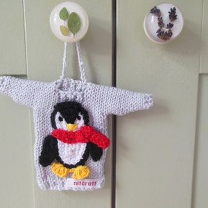 May include: A light grey knitted sweater ornament featuring a crocheted penguin appliqué. The penguin has a black and white body, a yellow beak and feet, and a bright red scarf. A small white tag at the bottom hem displays the text nitcraft.