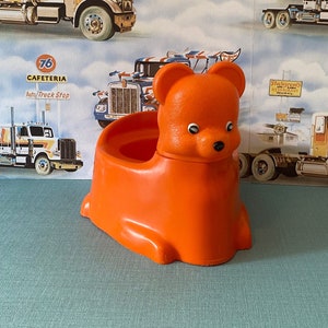 Puede incluir: Asiento de orinal para niños de plástico naranja con forma de oso con ojos negros y nariz negra. El orinal está sentado sobre una superficie azul con un fondo de papel tapiz con un patrón de camiones de plataforma.