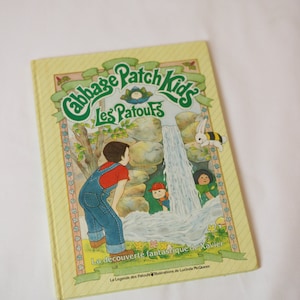 Peut inclure: Un livre jaune avec une illustration verte et blanche de personnages Cabbage Patch Kids. Le titre du livre est "Cabbage Patch Kids Les Patoufs" et le texte sur la couverture est "La découverte fantastique de Xavier" et "La Legende des Patoufs Illustrations de Lucinda McQueen".