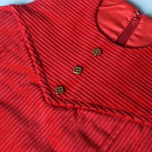 May include: Red corduroy dress with a high neckline and three gold buttons. The dress has a ribbed texture and a fitted silhouette.