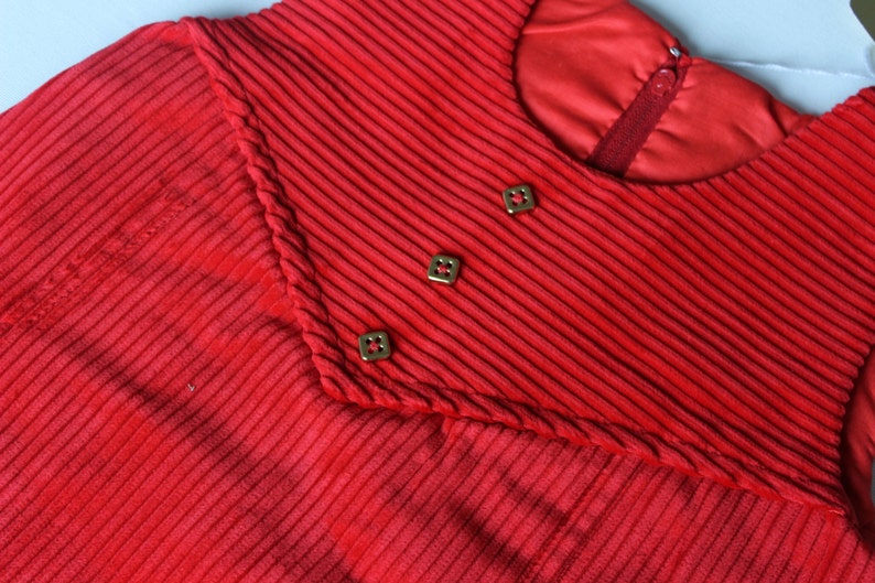 May include: Red corduroy dress with a high neckline and three gold buttons. The dress has a ribbed texture and a fitted silhouette.