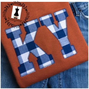 May include: A brown sweatshirt with a blue and white checkered "KY" design. The sweatshirt is folded and lying on a pair of blue jeans.