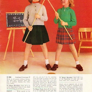May include: Two girls wearing knitted sweaters and skirts are holding wooden sticks. The girl on the left is wearing a red sweater and a black skirt. The girl on the right is wearing a green sweater and a red plaid skirt. The girls are standing in front of a red background with a small wooden easel and a chair.