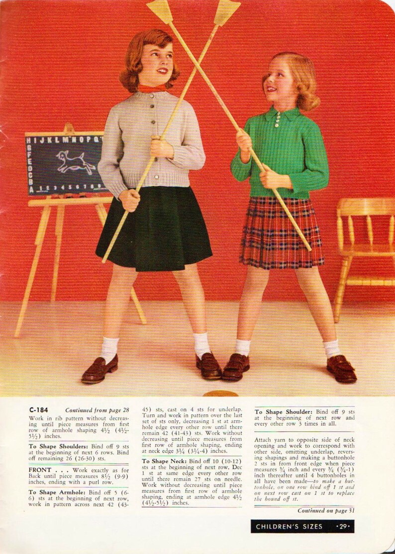 May include: Two girls wearing knitted sweaters and skirts are holding wooden sticks. The girl on the left is wearing a red sweater and a black skirt. The girl on the right is wearing a green sweater and a red plaid skirt. The girls are standing in front of a red background with a small wooden easel and a chair.