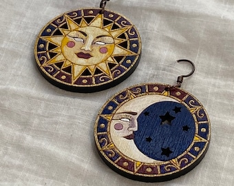 May include: A pair of round wooden earrings. One earring features a smiling sun with yellow, purple, and gold accents. The other earring depicts a crescent moon with a blue night sky and stars. Both earrings have a dark brown border and copper-colored ear wires.