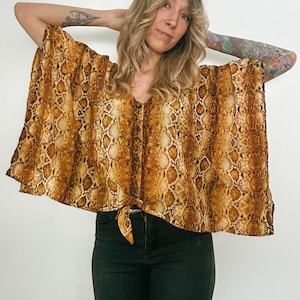 May include: A snakeskin-print top with a V-neck and button-down front. The top is tied at the waist and has wide, flowing sleeves. The colour palette includes shades of brown, tan, and gold. The top is paired with black jeans.