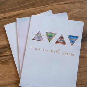 May include: A stack of notebooks with a white cover and the text "I am one with nature" in gold script. The cover features four colorful triangle designs. The notebooks are on a wooden surface.