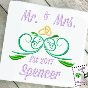 May include: Embroidered design featuring a green and white floral motif with two gold rings on top. The text "Mr. & Mrs. Est 2017 Spencer" is embroidered in purple below the design.