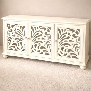 Puede incluir: Aparador de madera color crema con tres puertas. Cada puerta presenta un diseño calado decorativo. El mueble tiene una parte superior rectangular, pequeños pies redondos y pomos plateados. El aparador está sobre una alfombra clara.