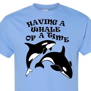 Puede incluir: Camiseta azul claro con un gr&aacute;fico negro de dos orcas y el texto "Having a Whale of a Time" en una fuente retro.