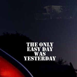 May include: A car window with the text "THE ONLY EASY DAY WAS YESTERDAY" in white, bold font. The image has a dark, moody aesthetic with a blurred background and a red taillight visible.