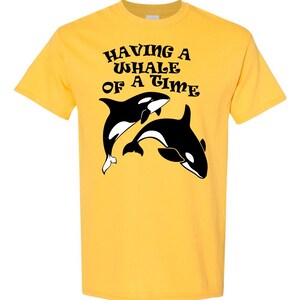 Puede incluir: Camiseta amarilla con un gr&aacute;fico negro de dos orcas y el texto "HAVING A WHALE OF A TIME" en una fuente retro.