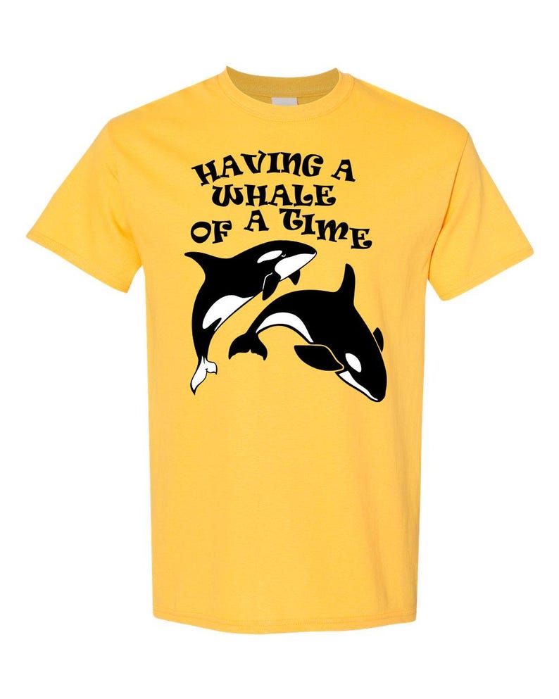 Puede incluir: Camiseta amarilla con un gr&aacute;fico negro de dos orcas y el texto "HAVING A WHALE OF A TIME" en una fuente retro.