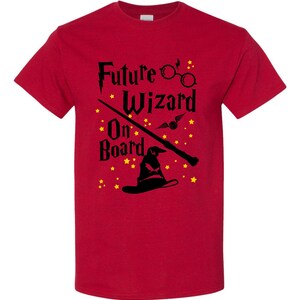May include: A red t-shirt with the words "Future Wizard On Board" in black, along with illustrations of glasses, a wand, a sorting hat, and stars. The shirt is made of a soft material.