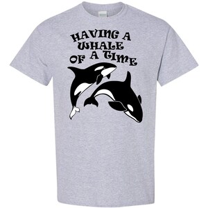 Puede incluir: Una camiseta gris con un gr&aacute;fico negro de dos orcas y el texto "HAVING A WHALE OF A TIME" en una fuente retro.