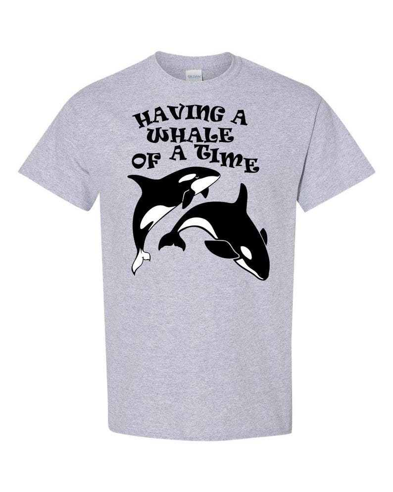 Puede incluir: Una camiseta gris con un gr&aacute;fico negro de dos orcas y el texto "HAVING A WHALE OF A TIME" en una fuente retro.