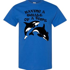 Puede incluir: Una camiseta azul real con un gr&aacute;fico en blanco y negro de dos orcas. El texto "HAVING A WHALE OF A TIME" est&aacute; impreso en negro en la camiseta.