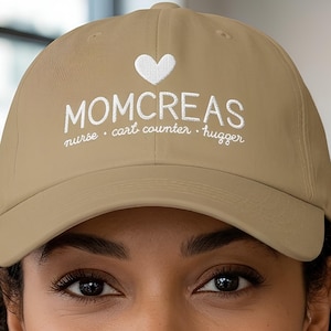 May include: A tan baseball cap featuring white embroidered text that reads MOMCREAS with a small heart icon above it and the words nurse, cart counter, hugger written in a smaller script font underneath the main text on the front panel.