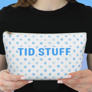 May include: White cosmetic bag with a blue polka dot pattern and the text "TID STUFF" in blue. The bag has a gold zip and is held by a person wearing a black top.