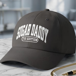May include: A dark gray baseball cap featuring white embroidered text on the front that reads SUGAR DADDY TID EDITION in a bold, arched collegiate font. The cap has a structured crown, a curved brim, and visible stitching details across the panels.