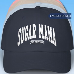 May include: A navy blue baseball cap featuring white embroidered text that reads SUGAR MAMA with T1D EDITION underneath in a smaller oval frame. The cap has a structured crown and a curved brim, with a small blue tag labeled EMBROIDERED.