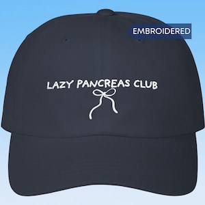 May include: A navy blue baseball cap featuring white embroidered text that reads LAZY PANCREAS CLUB above a white embroidered ribbon bow graphic. A small blue rectangular tag in the upper right corner contains the white text EMBROIDERED.