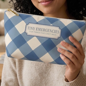 May include: A blue and cream gingham patterned zippered pouch featuring a rectangular label with the text TINY EMERGENCIES FOR THINGS THAT CAN'T WAIT. The pouch has a gold-toned metal zipper pull and a structured, fabric-like exterior finish.