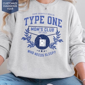 May include: A light grey sweatshirt with blue text. The text reads "TYPE ONE MOM'S CLUB EST. 2023" with a juice box graphic. The words "WHO NEEDS SLEEP?" are below the graphic. The words "CUSTOMIZE DIAGNOSIS YEAR" are in a blue box.