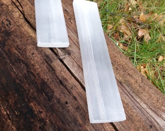 May include: Two white selenite sticks, also known as satin spar, are placed on a rough, brown wooden log. The sticks are smooth and have a natural, polished finish.
