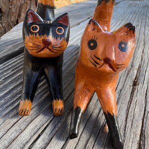 May include: Two wooden cat figurines. One cat is black with orange accents, the other is orange with black accents. Both have large eyes and are standing on four legs. The cats are hand-carved and painted.