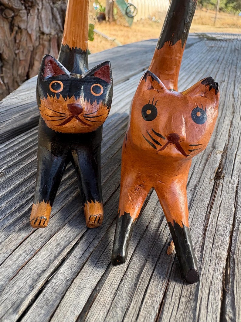 May include: Two wooden cat figurines. One cat is black with orange accents, the other is orange with black accents. Both have large eyes and are standing on four legs. The cats are hand-carved and painted.