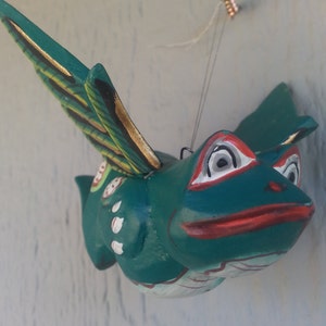 May include: A green wooden frog with gold accents and red and white details, flying with its wings outstretched. The frog is hanging from a string attached to a screw in the wall.