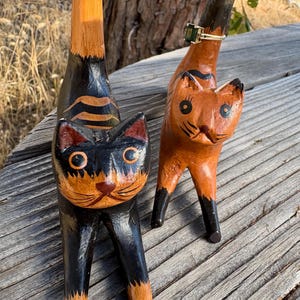 May include: Two wooden cat figurines, one black and orange, the other orange. The black cat has orange accents and stripes, while the orange cat has black stripes. Both have detailed facial features and are standing on a wooden surface.