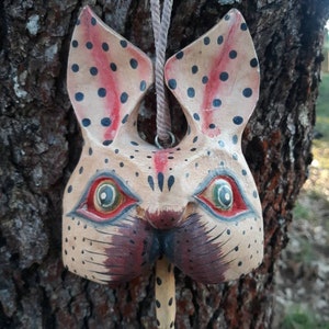 May include: A wooden mask of a rabbit with black and red polka dots, green and yellow eyes, and a long, brown, spotted nose. The mask is hanging from a brown rope.