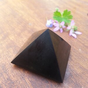May include: Black obsidian pyramid crystal on a wooden surface. The pyramid is smooth and polished.